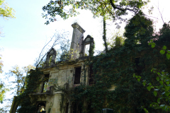 The image is a photograph of a partially ruined, two-story stone building, likely a castle or a large house, with a prominent arched window on the upper floor. The building is covered in dense, green ivy, which obscures much of the stonework and adds a sense of age and abandonment. The arched window is partially intact, with its pointed top still visible, and the lower part of the window is filled with ivy. The building's walls are made of large, irregularly shaped stones, giving it a medieval appearance. The roof is missing, exposing the interior to the elements. The ground floor has a few small, rectangular windows, also covered in ivy. The building is surrounded by tall, mature trees, with their leaves forming a natural canopy. The sky is visible in the top left corner, showing a clear blue sky with a few scattered clouds. The overall scene is one of serene decay, with the building's stone structure contrasting sharply with the vibrant green of the ivy and the trees. The image is taken from a low angle, emphasizing the height and grandeur of the building, despite its ruined state. The photograph captures the essence of a historical structure reclaimed by nature.