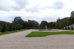 The image is a high-resolution photograph of a grand, symmetrical garden path leading to a distant fountain. The path is composed of light-colored gravel, bordered by meticulously trimmed, dark green hedges on both sides. The hedges are arranged in a geometric pattern, creating a sense of order and symmetry. In the foreground, there is a small, circular bed of vibrant green grass, adding a touch of natural softness to the otherwise structured scene. The path extends into the distance, leading the eye towards a tall, elegant fountain with a slender, white column topped by a statue, which is partially obscured by the distance. The fountain is surrounded by a circular pool of water, reflecting the sky and the surrounding greenery. The background consists of a dense, mixed forest of deciduous trees, their leaves displaying a mix of green and autumnal hues, suggesting a transition between seasons. The sky above is partly cloudy, with soft, white clouds scattered across a blue sky, adding a sense of tranquility to the scene. The overall mood of the image is serene and orderly, with a strong emphasis on symmetry and natural beauty. The photograph captures the essence of a well-maintained, classical garden, evoking a sense of peace and serenity. The image is taken from a low angle, emphasizing the length and grandeur of the path and the fountain. The photograph is clear and sharp, with excellent detail and color accuracy. The image is likely taken during the daytime, as indicated by the natural light and the clear sky. The photograph is devoid of human presence, focusing solely on the natural and man-made elements of the garden. The image is a testament to the beauty of classical garden design, with its emphasis on symmetry, order, and natural beauty. The photograph is likely taken with a high-quality digital camera, as indicated by the sharpness and clarity of the image. The image is a digital file, with a resolution of 300 dpi, suitable for printing and display.