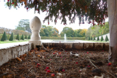 The image is a high-resolution photograph capturing a serene garden scene. In the foreground, there is a low, white stone wall with a smooth, slightly curved top, partially covered with red and orange maple leaves. The wall leads the eye towards a central focal point: a tall, white stone pedestal with a spherical top, standing on a circular base. The pedestal is positioned near the edge of a rectangular pond, which reflects the sky and surrounding greenery. The pond is bordered by a stone edge, and a small fountain is visible in the center, spraying water upwards in a gentle arc. The background consists of a lush, green garden with various trees and shrubs, including a prominent maple tree with vibrant red leaves on the right side of the image. The sky is overcast, with a soft, diffused light illuminating the scene. The overall mood is tranquil and slightly melancholic, with the autumnal colors adding a touch of warmth and decay. The photograph is taken from a low angle, emphasizing the height of the pedestal and the expanse of the garden. The image is rich in detail, with a clear focus on the textures and colors of the natural elements. The composition is balanced, with the pedestal and pond serving as the central point of interest, framed by the stone wall and the garden's greenery. The photograph is likely taken during the autumn season, as indicated by the falling leaves and the overall color palette. The image is devoid of human presence, emphasizing the natural beauty of the garden. The photograph is a blend of natural and man-made elements, with the stone structures and the garden's greenery coexisting harmoniously. The image is a testament to the photographer's skill in capturing the essence of a serene garden landscape. The photograph is likely taken with a high-quality digital camera, as indicated by the sharpness and clarity of the image.