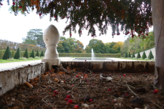 The image is a high-resolution photograph capturing a serene garden scene. In the foreground, there is a stone wall with a rough texture, constructed from large, irregularly shaped stones. The wall is slightly curved, adding a sense of depth and perspective to the image. On top of the wall, there is a small, ornate stone statue of a bird, possibly a pigeon or a dove, perched on a pedestal. The statue is detailed, with a smooth, glossy finish, contrasting with the rough texture of the wall.Beyond the wall, the garden is lush and well-maintained, with a variety of trees and shrubs. The trees are a mix of deciduous and coniferous types, with some displaying autumn foliage in shades of red, orange, and yellow. The shrubs are neatly trimmed, adding to the overall orderliness of the garden. In the distance, there is a large, rectangular pond with a fountain at its center, spraying water upwards in a graceful arc. The pond is surrounded by a stone edge, and the water appears calm and still, reflecting the sky above.The sky is overcast, with a uniform gray tone, suggesting a cloudy day. The overall mood of the image is tranquil and peaceful, with a sense of natural beauty and serenity. The photograph is taken from a low angle, looking up at the wall and the statue, emphasizing the height and grandeur of the scene. The image is rich in detail and texture, from the rough stone wall to the smooth, glossy statue, and the lush greenery of the garden. The photograph is likely taken during the daytime, as indicated by the natural light and the clear visibility of the scene. The image does not contain any text or human figures, focusing solely on the natural elements of the garden. The photograph is a testament to the beauty of natural landscapes and the artistry of garden design.