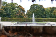 The image is a high-resolution photograph capturing a serene garden scene. In the foreground, there is a rectangular stone pool with clear water, and a central water jet creating a vertical stream. The pool is surrounded by a low stone wall, and the water is calm, reflecting the sky and surrounding greenery. The garden is lush with various types of trees and shrubs, including tall, dense evergreens and deciduous trees with a mix of green and autumnal foliage. The sky is partly cloudy, with soft, diffused light illuminating the scene. The overall atmosphere is tranquil and inviting, with a sense of natural beauty and serenity. The photograph is taken from a slightly elevated perspective, emphasizing the pool and the garden's layout. The image is rich in detail, capturing the textures of the stone, the smoothness of the water, and the varying shades of green in the foliage. The composition is balanced, with the pool and water jet as the focal point, framed by the garden's natural elements. The photograph is likely taken during the daytime, as indicated by the natural light and the clear sky. The image does not contain any human figures or man-made structures, emphasizing the natural beauty of the garden. The photograph is likely taken with a digital camera, given its high resolution and clarity. The image is devoid of any text or symbols, focusing solely on the natural elements. The overall mood is peaceful and contemplative, inviting the viewer to appreciate the tranquility of the garden setting. The photograph is a testament to the beauty of natural landscapes and the calming effect of water features in garden design. The image is likely taken in a private garden or a public park, given its well-maintained and serene atmosphere. The photograph is a realistic representation of the scene, capturing the essence of the garden's natural beauty. The image is devoid of any artistic manipulation, focusing solely on the natural elements and their interplay.